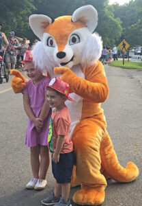 The fox mascot stops to take a picture with a young boy and girl at a park event.