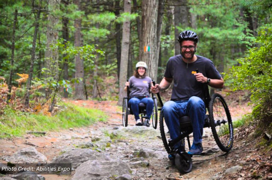 Image of two adults on all terrain wheelchairs hiking on a nature trail in a wooded area